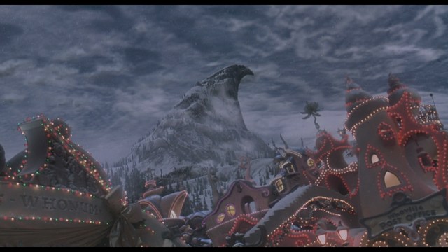 A man with large white wings stands in a room, wearing a dark suit, with a serious expression on his face. Bookshelves and curtains are visible in the background.