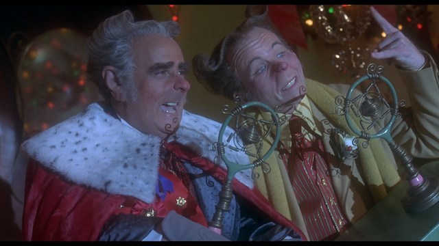 A man with short hair wearing a brown jacket and purple shirt, speaking animatedly in front of a colorful restaurant featuring a cartoon cow logo in the background.