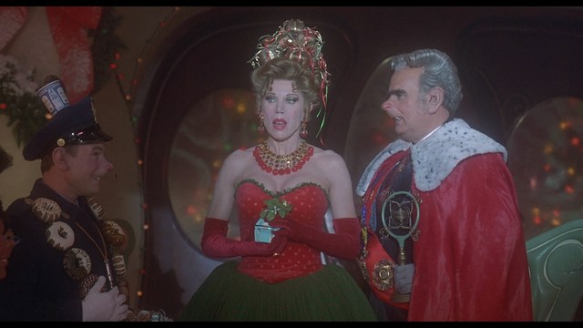 A bronze trophy figure of a cartoonish character with arms outstretched, positioned on a wooden table in a corporate setting, with a large window showcasing a mansion in the background.