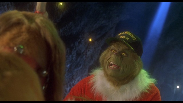 A scene from a movie featuring a man with short black hair and a dark jacket, sitting at a table with a surprised expression, holding a beverage, in a dimly lit indoor setting.