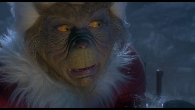 A close-up shot of a woman with long dark hair, wearing a maroon top and earrings, sitting in a dimly lit setting with red stools and decorative artwork in the background.