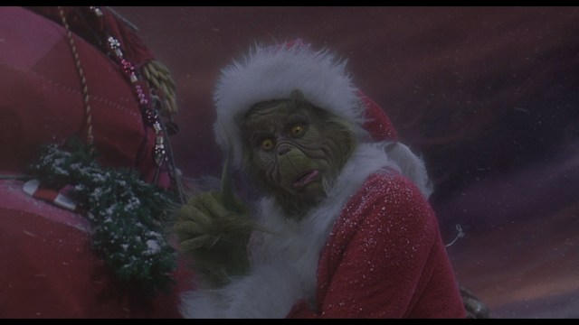 Close-up shot of a young man with short hair, wearing a gray hoodie, looking over his shoulder with a contemplative expression, while another person is asleep in the background.