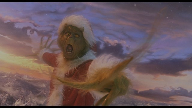 Two male characters in a cozy indoor setting, one with long hair and a beanie, smirking while holding a small object, and the other with a beard and cap, looking surprised. A colorful background with posters can be seen.