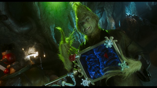 A woman with a pensive expression rests her chin on her hand, sitting at a table with kitchen appliances and items in the background.