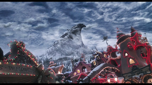 A man with large, feathered angel wings stands in a room, wearing a black suit and looking directly at the viewer.