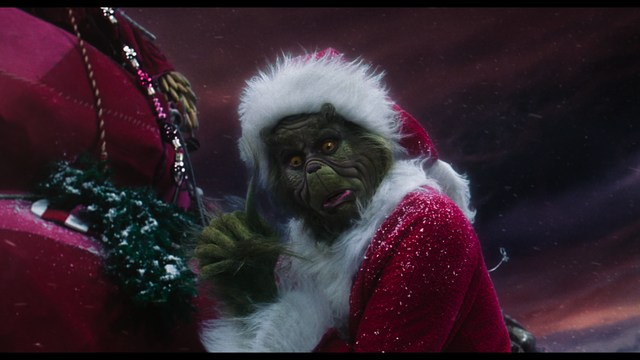 A close-up shot of a young man with short hair and a gray sweatshirt, looking intently at the camera, with a woman sleeping beside him in the background.