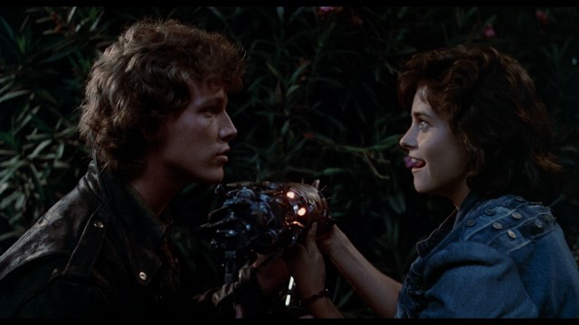 Two young men are looking at something off-camera, standing inside a building with bright lighting and seating areas visible in the background.