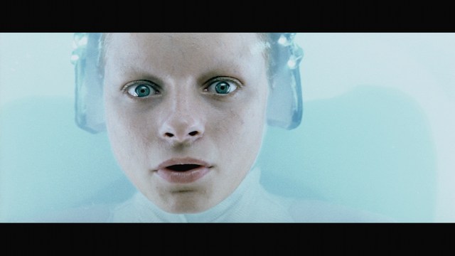 A man stands in front of an amusement arcade with a mural featuring a playful snowman and signs advertising games like skeeball, in a beach setting.