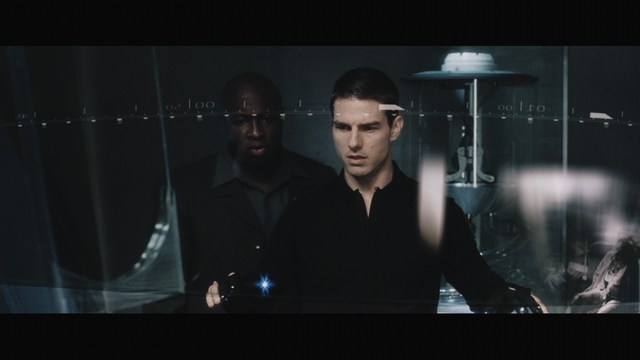 A woman with long dark hair, wearing a brown sweater, gestures with one hand while speaking in a warmly lit room with blinds and colorful artwork in the background.