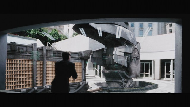 Close-up of a man with a serious expression, seated in a dimly lit setting, with another person in the background appearing to be seated at a table.