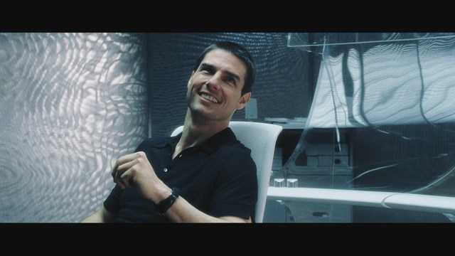 A scene from a movie featuring a man with short black hair and a dark jacket, sitting at a table with a surprised expression, holding a beverage, in a dimly lit indoor setting.