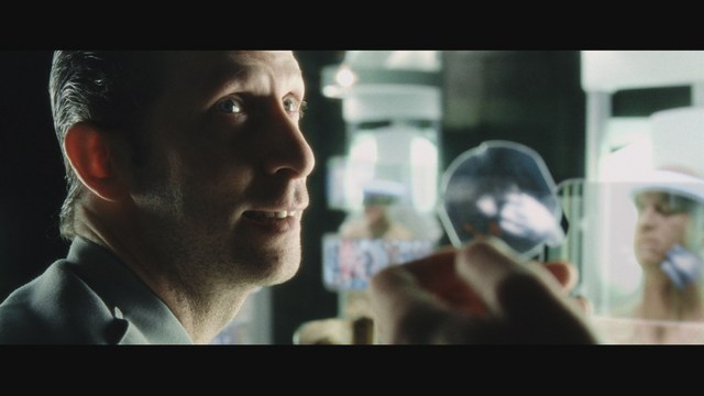 Close-up shot of a young man with short hair, wearing a gray hoodie, looking over his shoulder with a contemplative expression, while another person is asleep in the background.