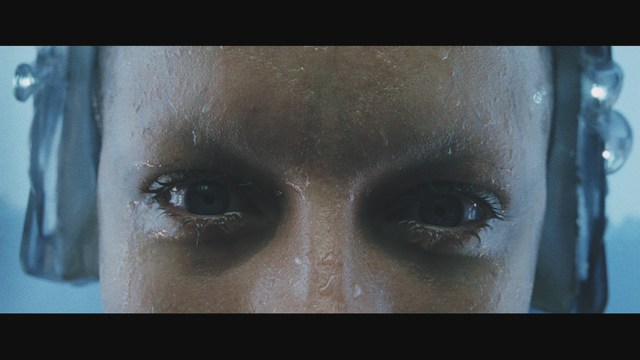 Two male characters in a cozy indoor setting, one with long hair and a beanie, smirking while holding a small object, and the other with a beard and cap, looking surprised. A colorful background with posters can be seen.