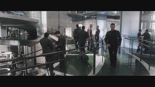 A scene from a movie featuring a man dressed in a bishop's robe speaking at a podium surrounded by microphones, with a woman in a red outfit smiling next to him, and an audience in the background.