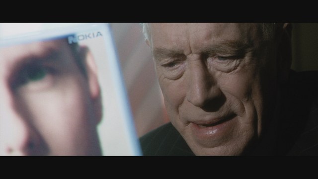 A man with long hair and a baseball cap appears shocked or surprised, sitting in a dimly lit room with wooden paneling and chairs in the background.