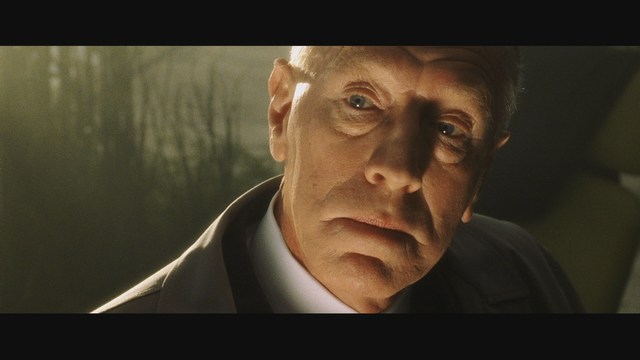 Close-up of a man with a serious expression, featuring details of his face and suit against a softly lit background.