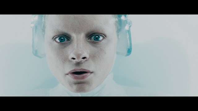 A still from a movie featuring a man standing on a boardwalk in front of an arcade with signs for Skeeball, saltwater taffy, and various attractions.