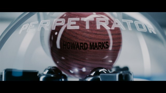 A man with a beard and mustache smiles warmly while standing on a boardwalk by the beach, with colorful flags overhead and a large building in the background.
