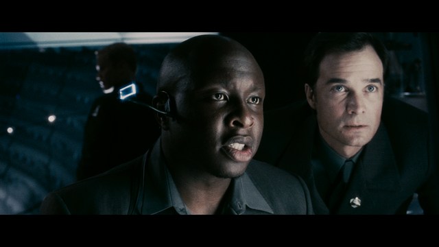 A man with large, feathered angel wings stands in a room, wearing a black suit and looking directly at the viewer.