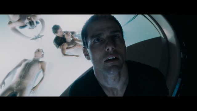 Two young men are looking at something off-camera, standing inside a building with bright lighting and seating areas visible in the background.