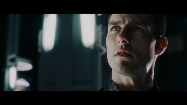 Close-up of a man with a serious expression, seated in a dimly lit area, with soft lighting from background lamps.