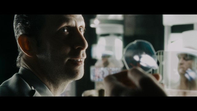 A close-up shot of a young man with short hair and a gray sweatshirt, looking intently at the camera, with a woman sleeping beside him in the background.