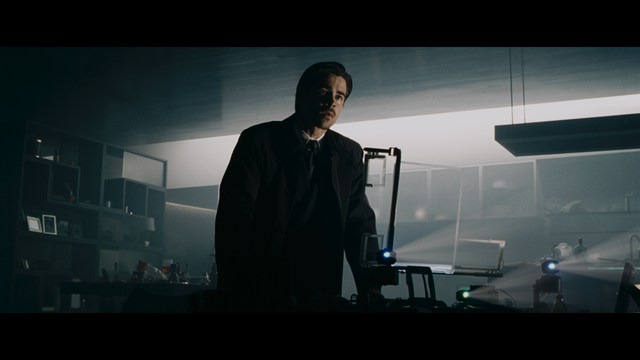 A woman seated at a restaurant table, thoughtfully resting her chin on her hand while reading a newspaper. Blurred figures of two people can be seen in the background.