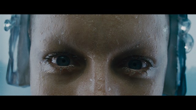 Two characters sitting at a table, one holding a small statue, with a surprised expression. The background features vibrant colors and patterns.