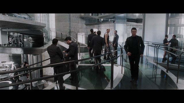 A scene featuring a man in a bishop's attire speaking at a podium surrounded by microphones, with a woman in a red dress beside him and a crowd of onlookers in the background.