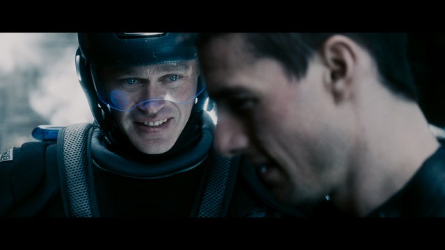 A scene from the film showing two male characters, one in a futuristic helmet and uniform, smiling while the other appears tense, conveying a sense of intrigue.