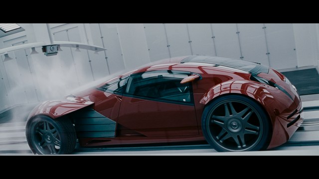 A sleek red futuristic car showcased in a high-tech environment, possibly a testing facility, with a modern design featuring large wheels and a distinctive silhouette.