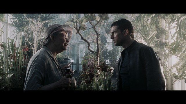 A scene from a film featuring two characters, one elderly woman holding a small object and speaking animatedly, and a young man listening intently, surrounded by lush greenery and plants in a greenhouse setting.