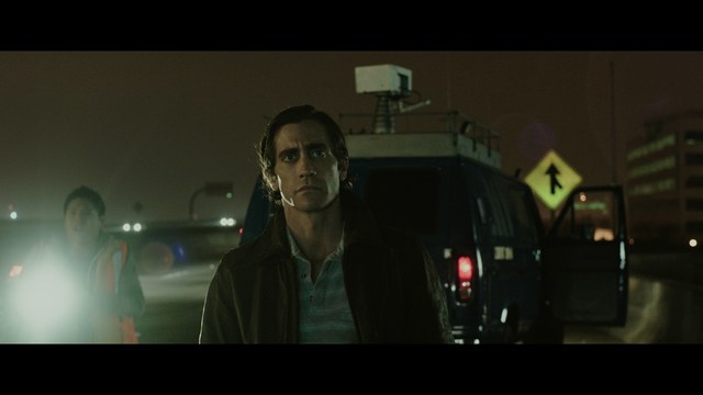 A serious-looking cardinal stands in front of a wooden backdrop with colorful posters related to Catholicism, holding a microphone at a press conference.