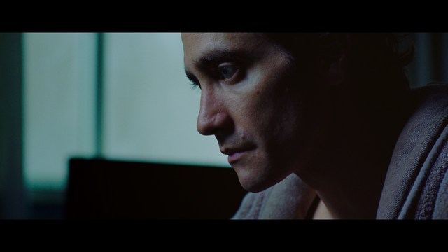 A woman with a pensive expression rests her chin on her hand, sitting at a table with kitchen appliances and items in the background.