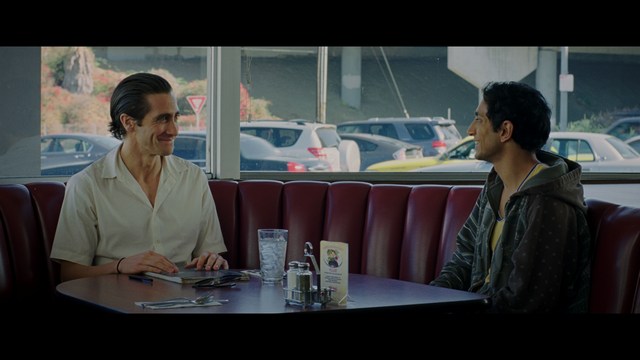 A man with large, feathered angel wings stands in a room, wearing a black suit and looking directly at the viewer.