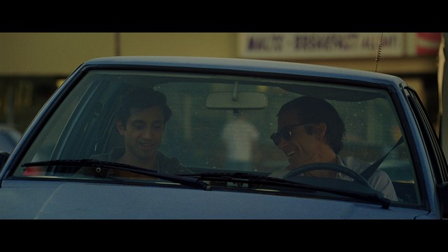 A close-up of a man with a concerned expression, with blurred figures in the background, set in a dimly lit interior.