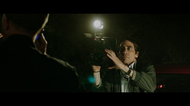 Two young men are looking at something off-camera, standing inside a building with bright lighting and seating areas visible in the background.