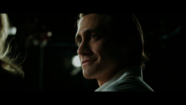 Close-up of a man with a serious expression, seated in a dimly lit area, with soft lighting from background lamps.