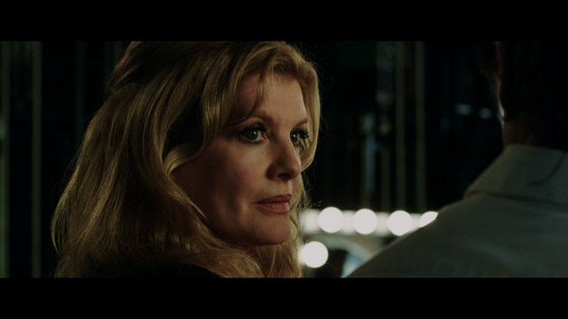 A close-up shot of a young man with short hair and a gray sweatshirt, looking intently at the camera, with a woman sleeping beside him in the background.