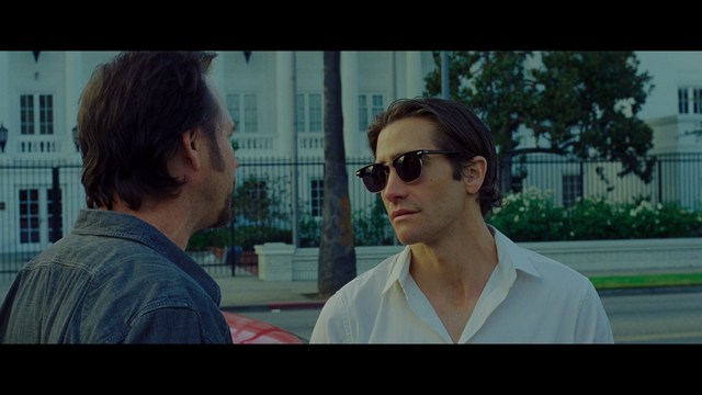 A woman seated at a restaurant table, thoughtfully resting her chin on her hand while reading a newspaper. Blurred figures of two people can be seen in the background.