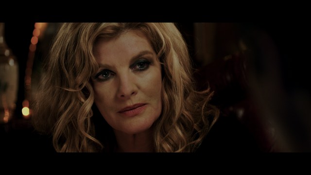 Two characters sitting at a table, one holding a small statue, with a surprised expression. The background features vibrant colors and patterns.