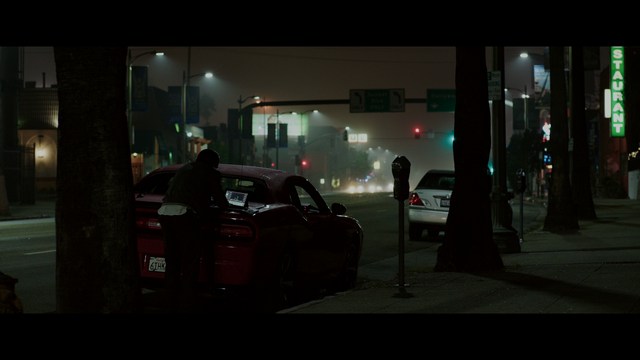A scene featuring a man in a bishop's attire speaking at a podium surrounded by microphones, with a woman in a red dress beside him and a crowd of onlookers in the background.