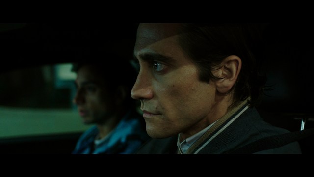 Close-up of an elderly woman in a wide-brimmed hat, with a serious expression. She is in an indoor setting with visible plants in the background.