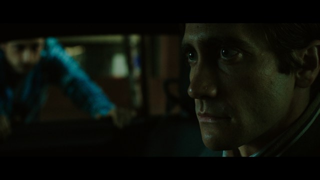 A scene from a film featuring two characters, one elderly woman holding a small object and speaking animatedly, and a young man listening intently, surrounded by lush greenery and plants in a greenhouse setting.