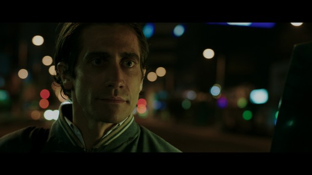 A scene from a film featuring two characters, one elderly woman holding a small object and speaking animatedly, and a young man listening intently, surrounded by lush greenery and plants in a greenhouse setting.