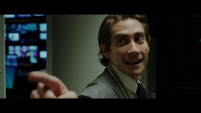 A scene from a film featuring two characters, one elderly woman holding a small object and speaking animatedly, and a young man listening intently, surrounded by lush greenery and plants in a greenhouse setting.