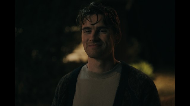 A close-up of a man with a concerned expression, with blurred figures in the background, set in a dimly lit interior.
