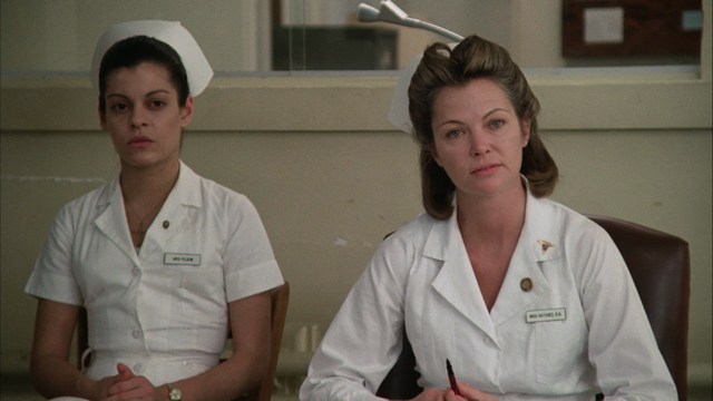 A woman with long dark hair, wearing a brown sweater, gestures with one hand while speaking in a warmly lit room with blinds and colorful artwork in the background.