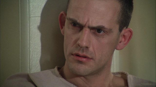 A man in a white suit and hat stands at the entrance of a house, smiling, with a suburban neighborhood visible in the background.