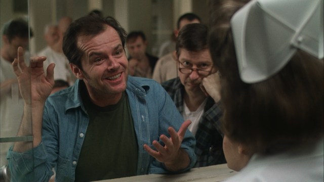 Close-up of a man with a serious expression, seated in a dimly lit setting, with another person in the background appearing to be seated at a table.